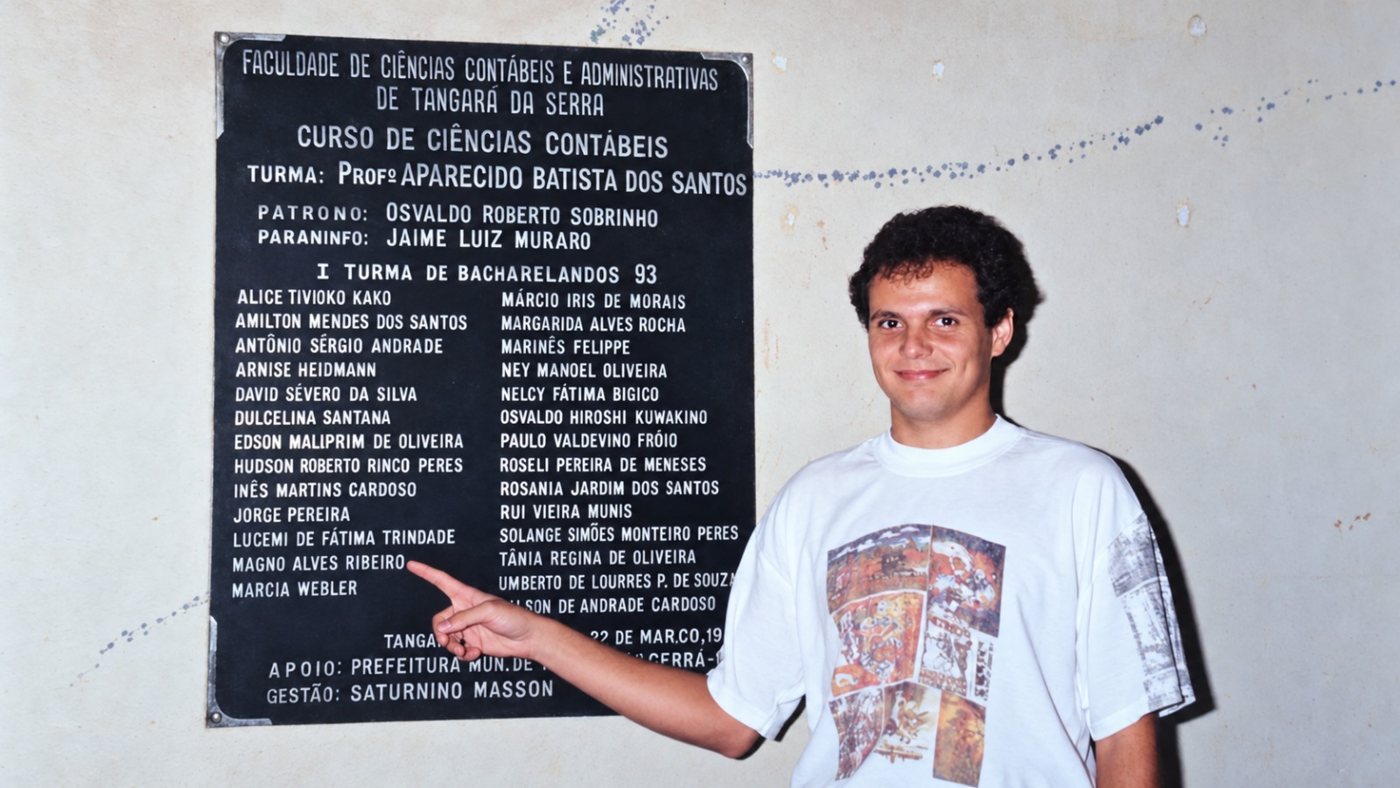 Magno jovem, de camiseta branca, apontando o próprio nome na placa da I Turma de Bacharelandos em Ciências Contábeis